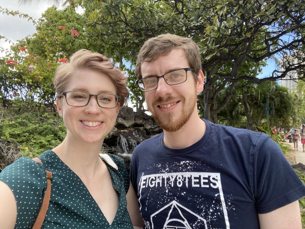 Amy and her partner Joel, a man with brown hair, stand in front of a row of tropical trees.