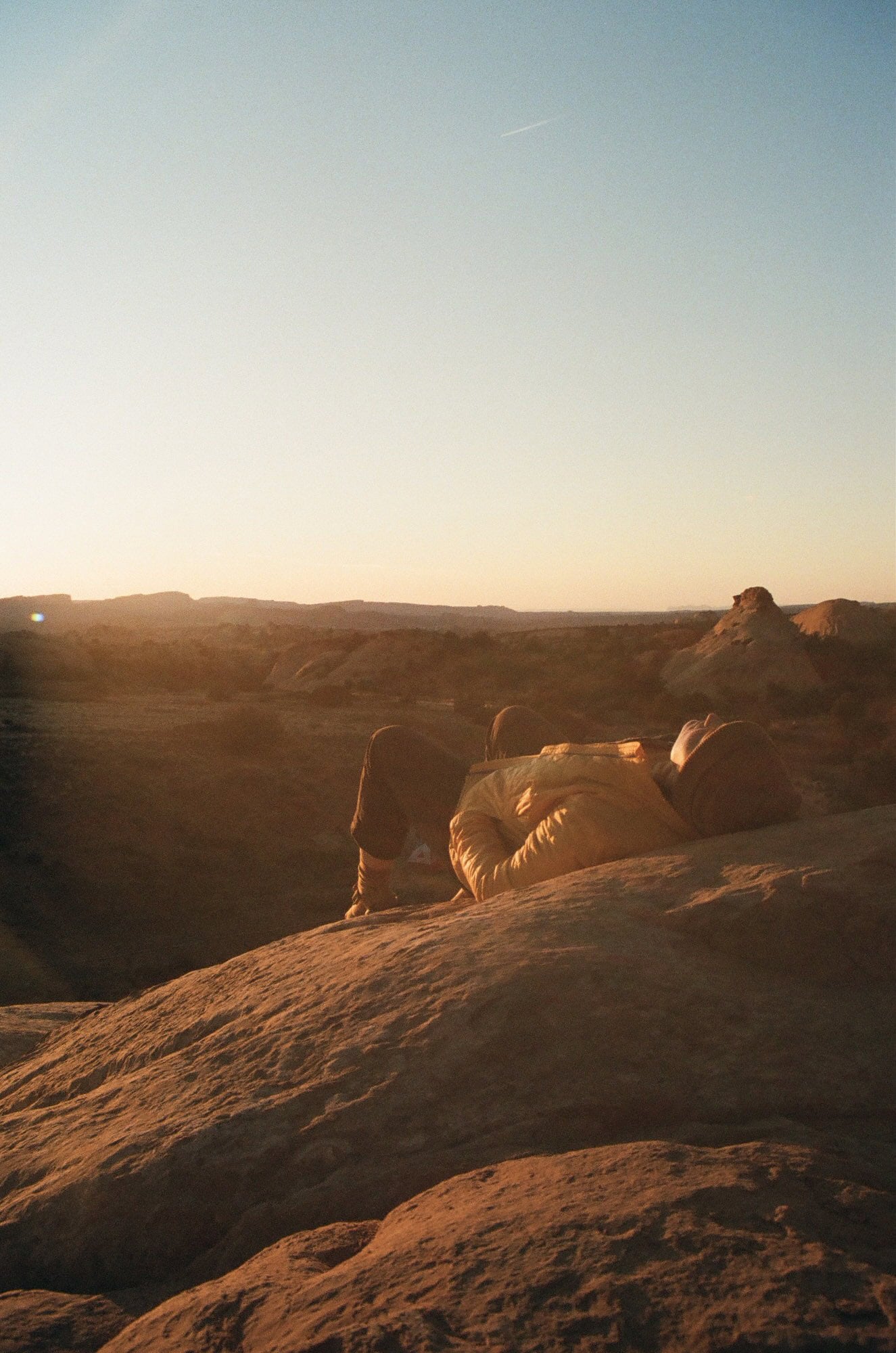 A person in a yellow jacket lies on a rocky surface at sunset, with a vast desert landscape in the background.