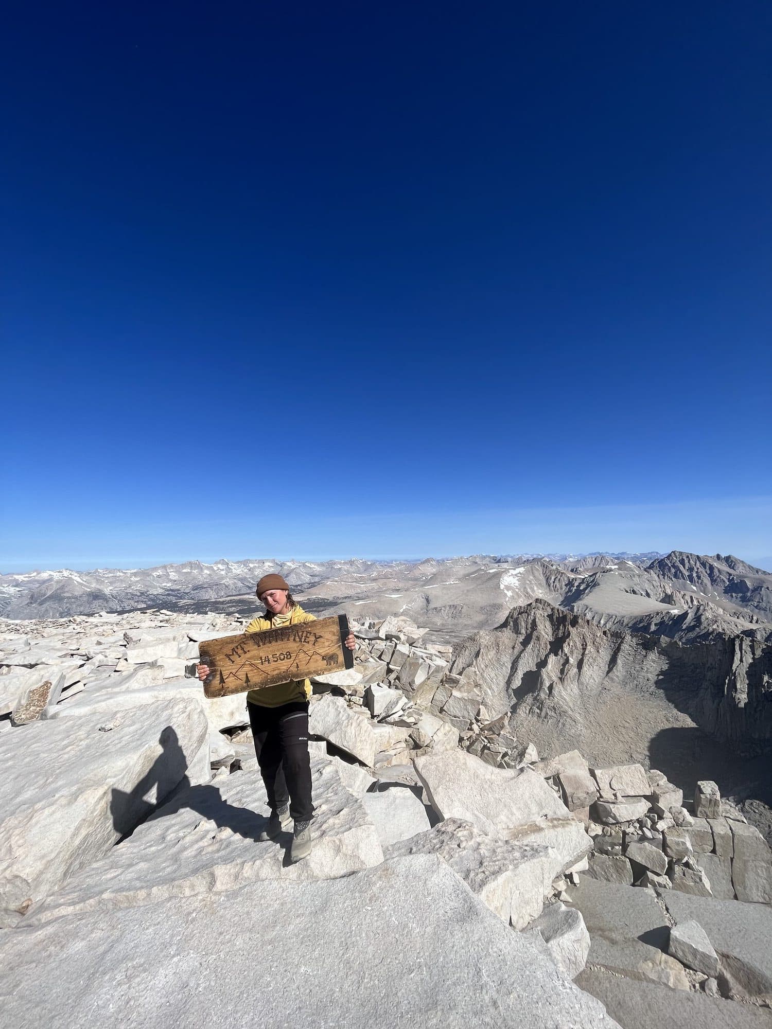 A person stands on a rocky mountain summit holding a sign that reads "Mt. Whitney, Elev. 14,508 ft," with a vast landscape and clear blue sky in the background.