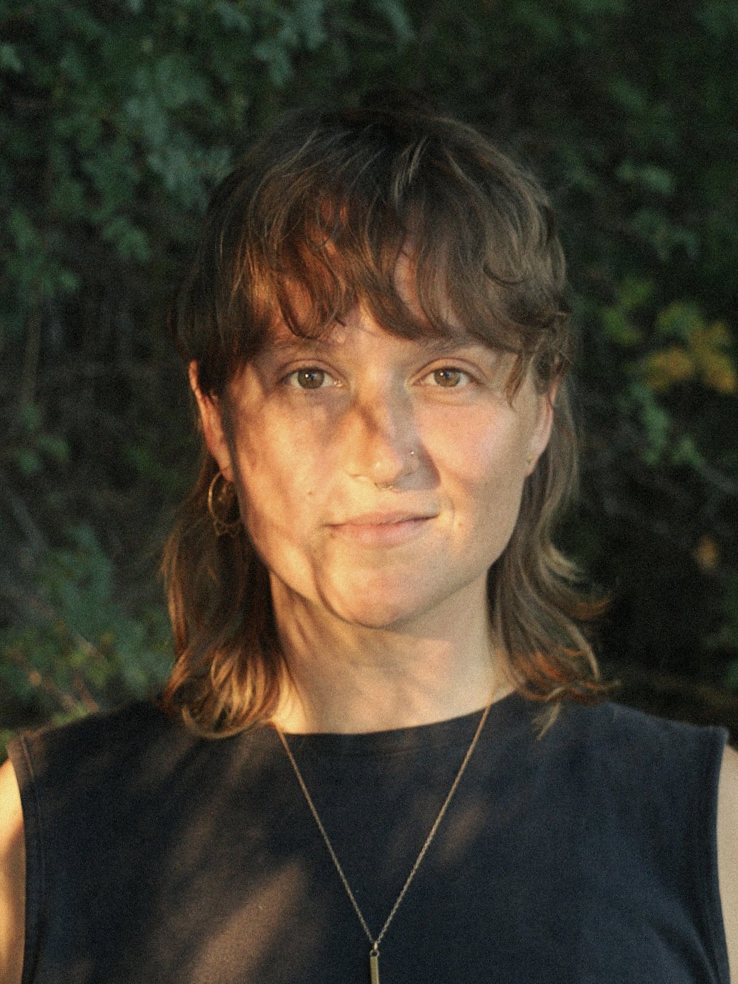 A person with short brown hair stands outdoors, wearing a sleeveless dark top and a necklace, with sunlight casting dappled shadows on their face. Green foliage is in the background.